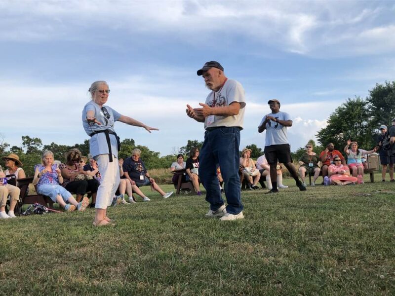 A group of people gathers on grass; two individuals engage in a playful interaction, some seated and observing, under a clear blue sky.