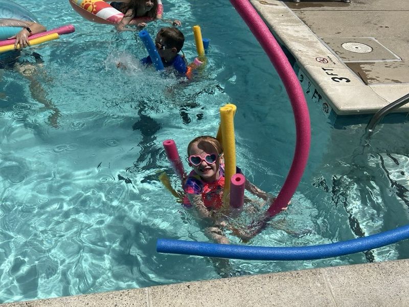 swim time Children and a person enjoy swimming in a pool with colorful float noodles and tubes, under sunny weather. Poolside measures 3 feet deep.