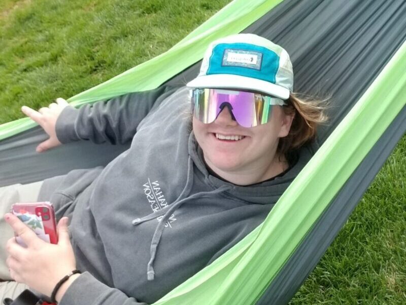 volunteer in eno A person wearing a cap and reflective sunglasses relaxes in a green hammock on grassy ground, holding a smartphone.