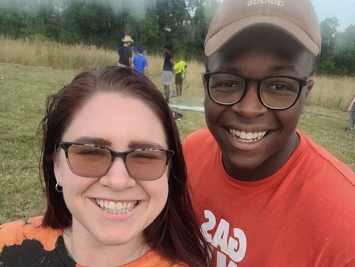 volunteering at claggett Two people smiling in a field, wearing casual shirts. Background includes other people and a grassy area with trees. No landmarks visible.