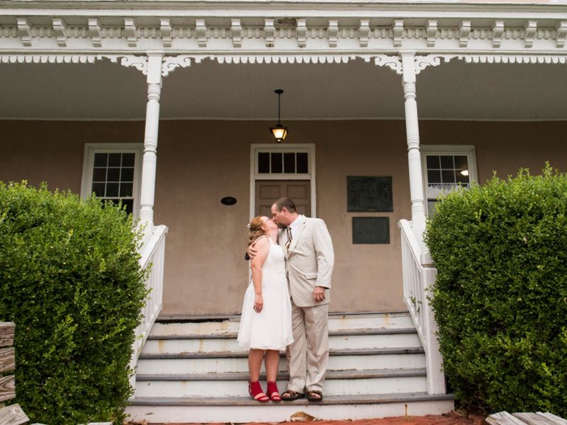 wedding-anniversary A couple kisses on the steps of a historic building's porch, featuring decorative pillars and trimmed hedges, under a vintage light.