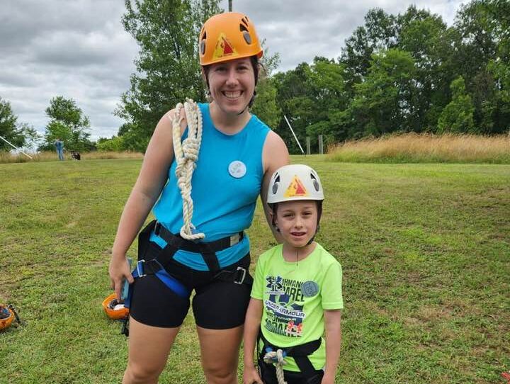 woman and girl in ropes gear Two people wearing helmets and harnesses stand on grass, ready for a climbing activity. Trees and a cloudy sky are in the background.