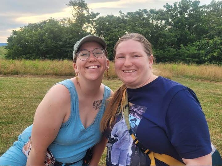 workers at claggett Two people smiling outdoors in a field with trees and hills in the background. A barbecue grill is nearby.