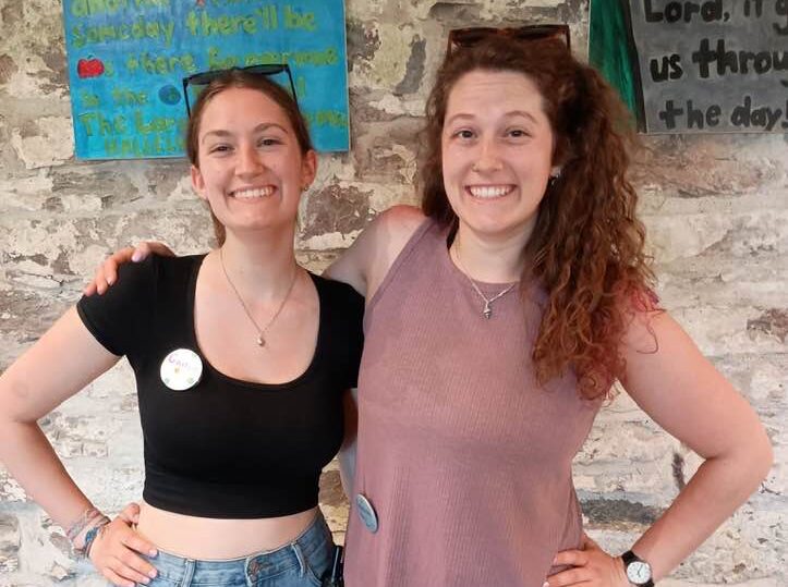 workers during camp Two people smiling in front of a stone wall with colorful signs featuring text. Casual attire and friendly demeanor are depicted.
