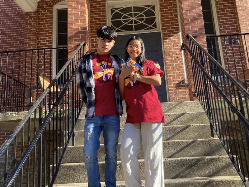 youth on stairs Two people in red shirts stand smiling on steps of a brick building with black railings, holding colorful packages.