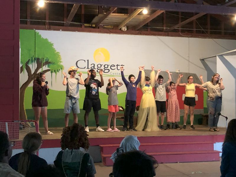 youth-play People on stage at Claggett Center, performing with raised arms. Audience watches a cheerful scene under stage lights.