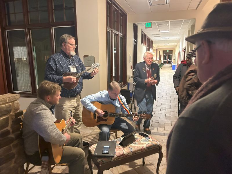 Mens retreat photo A group of people playing acoustic instruments in a hallway, surrounded by others. Warm indoor setting with tile flooring and wooden chairs.