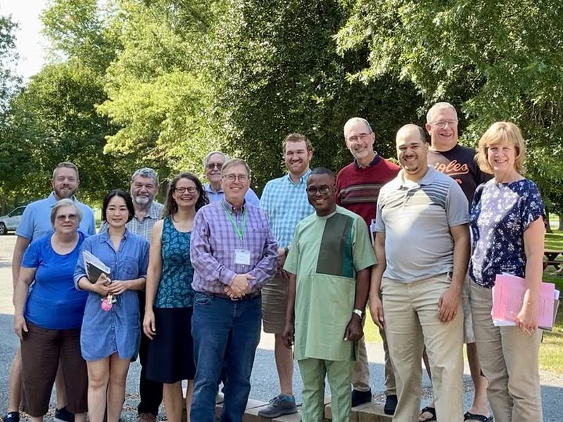 Musicians retreat photo A group of twelve people standing outdoors, smiling, with trees in the background on a sunny day. Casual attire, diverse age group.