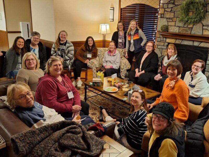 Women's Retreat A group of people sit together in a cozy room, sharing snacks around a coffee table near a stone fireplace decorated with a wreath.