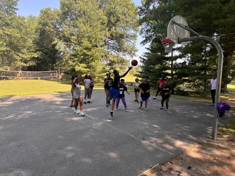 basketball at camp A group of people playing basketball outdoors on a sunny day, surrounded by trees and an adjacent volleyball net.