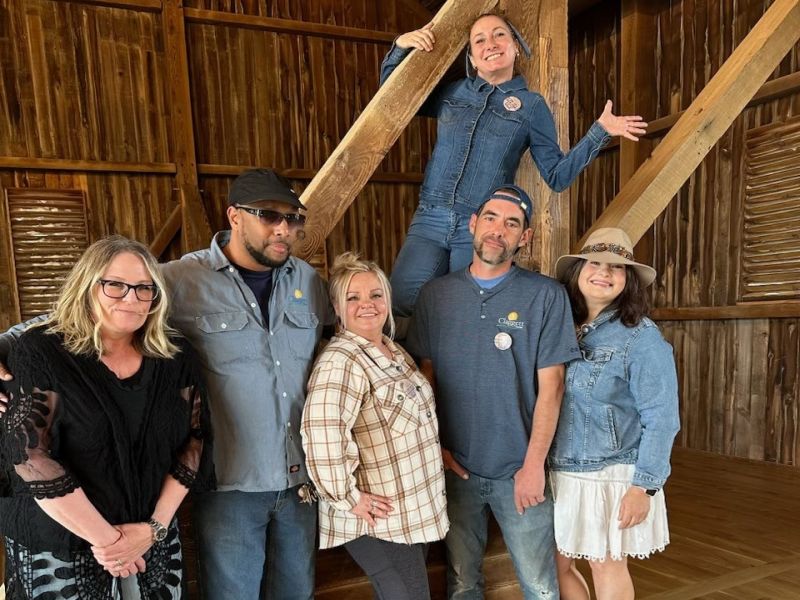 blue jean gala folks A group of six people in casual clothing pose inside a rustic wooden structure, with visible beams and planks in the background.