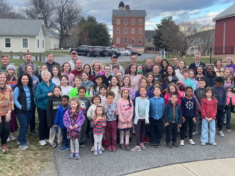camp 2024 A large group of people and children pose outdoors; a historic multi-story brick building is visible in the background.
