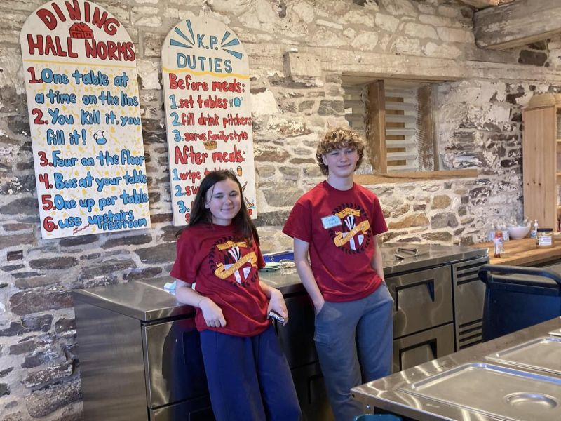 dining hall kids serving Two people stand in a rustic dining hall with stone walls, wearing matching red shirts. Instructional signs hang on the wall.