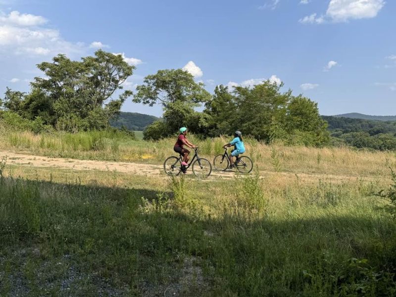 girls riding bikes Two people biking on a dirt path in a grassy landscape, surrounded by trees and hills under a clear blue sky.