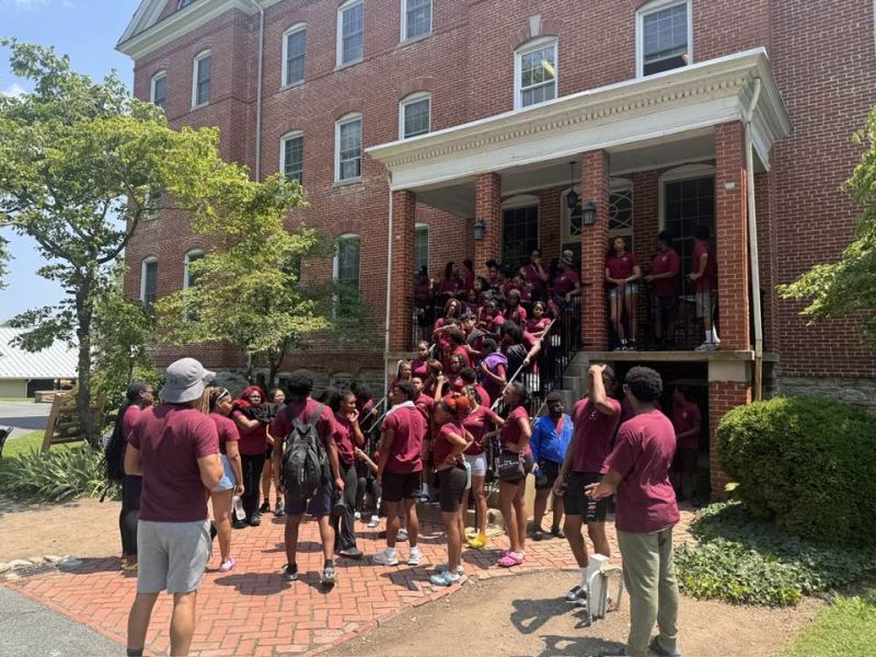 group retreat camp A group of people wearing maroon shirts gather outside a large brick building with a porch, surrounded by trees and a paved walkway.