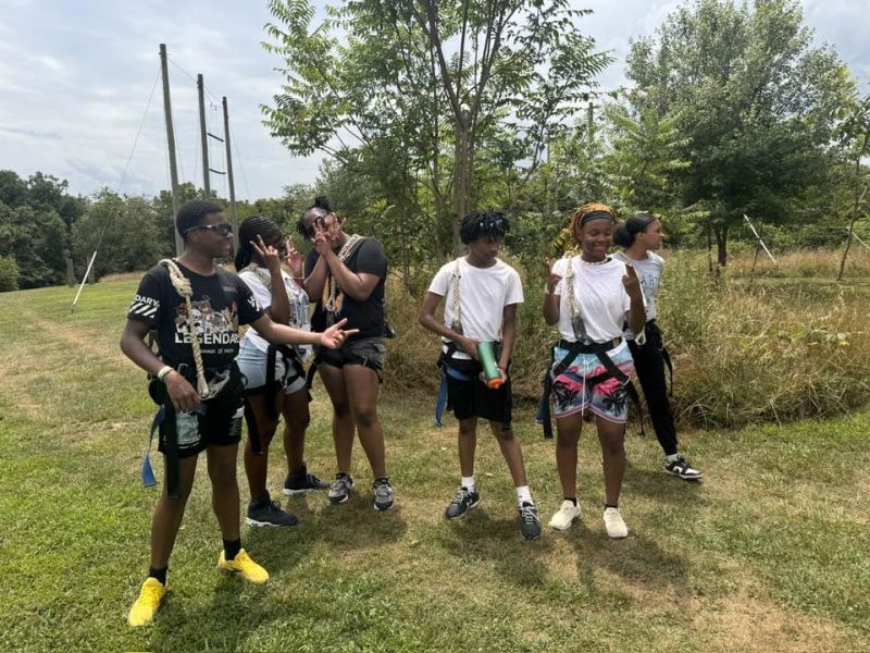 kids ready for course Six people wearing harnesses stand on a grassy field, surrounded by trees, appearing ready for an outdoor adventure activity.