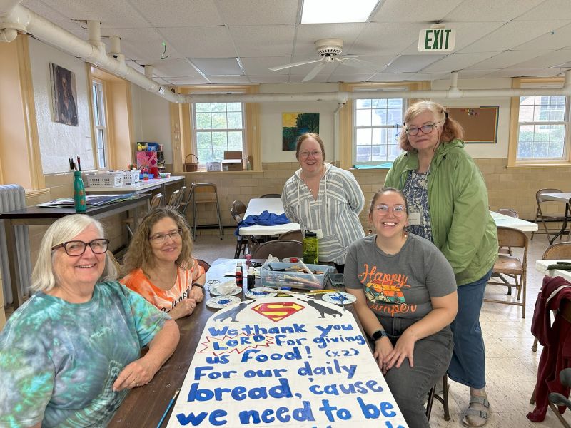 ladies at retreat Five people in a room with tables and chairs, smiling around a painted sign expressing gratitude for food.