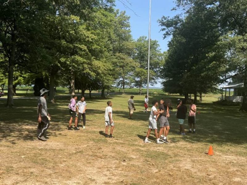 summer camp teather ball A group of people and children are gathered in a park with scattered trees and a tall flagpole.