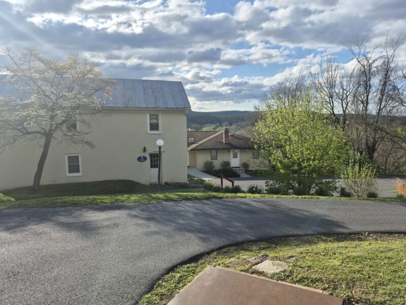 cottage A large, modern two-story building with beige paneling stands amidst green lawns, set against a cloudy sky. Metal chairs feature in the foreground.