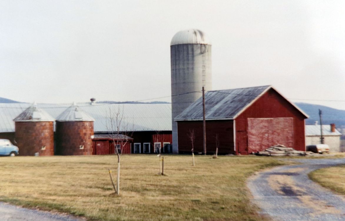 Farm scene with red barns, silos, and a winding path in a rural setting. Hilly landscape visible in the background. No landmarks identified.