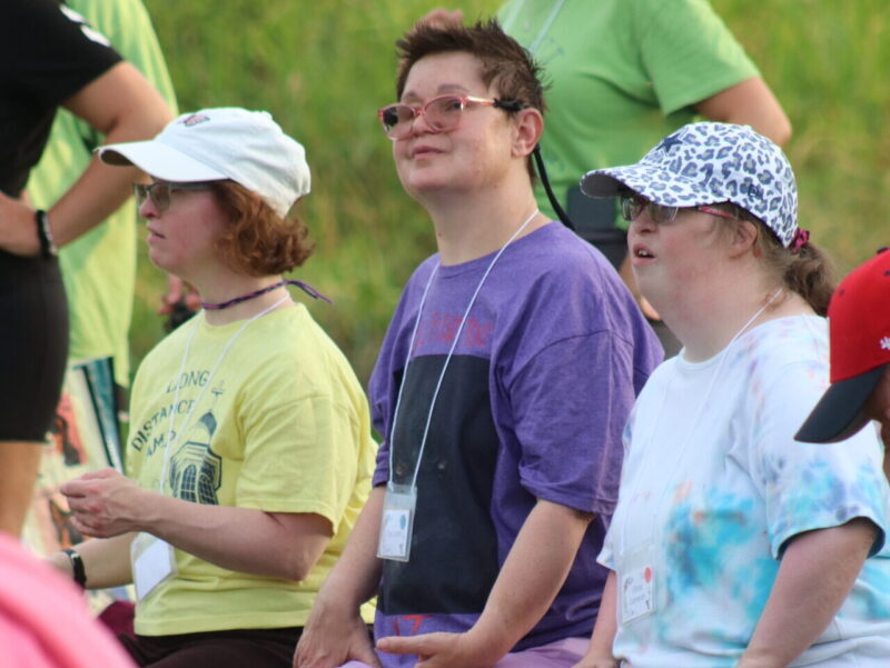 Three people at camp Three people sitting outdoors on a bench, wearing casual clothing and hats, surrounded by grass. They appear engaged in an event or gathering.