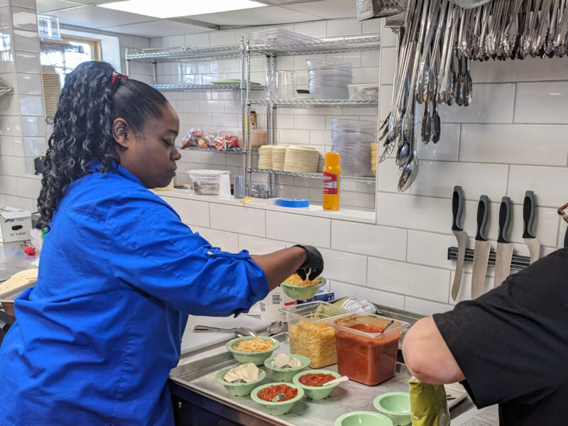 PXL_20240214_153107311 A person in a blue shirt prepares food in a commercial kitchen, surrounded by utensils, knives, and various ingredients on the counter.