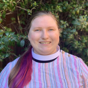 Sara Yoe A person smiles while wearing a multicolored striped shirt and white collar, standing in front of lush green foliage. No landmarks visible.