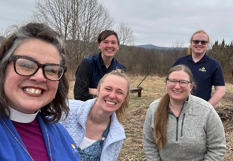 Five people smiling in an outdoor setting with barren trees and a cloudy sky, standing in an open field with distant hills.