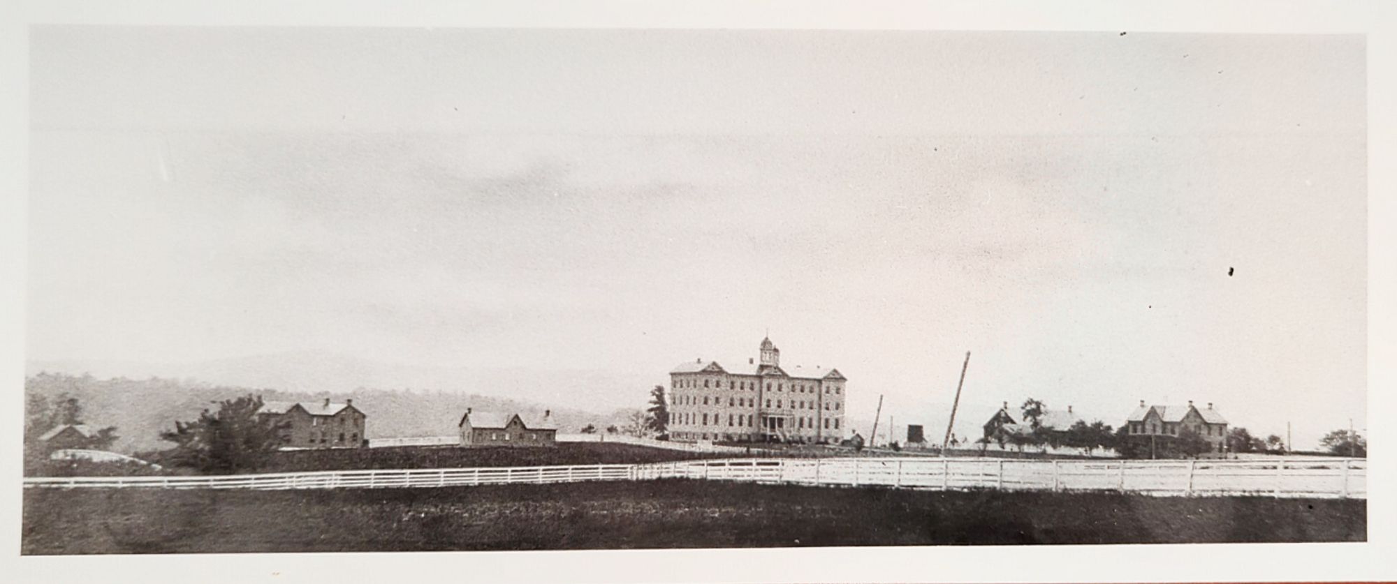 A historical black and white image shows a large, multi-story building with smaller structures surrounding it, all enclosed by a white wooden fence.