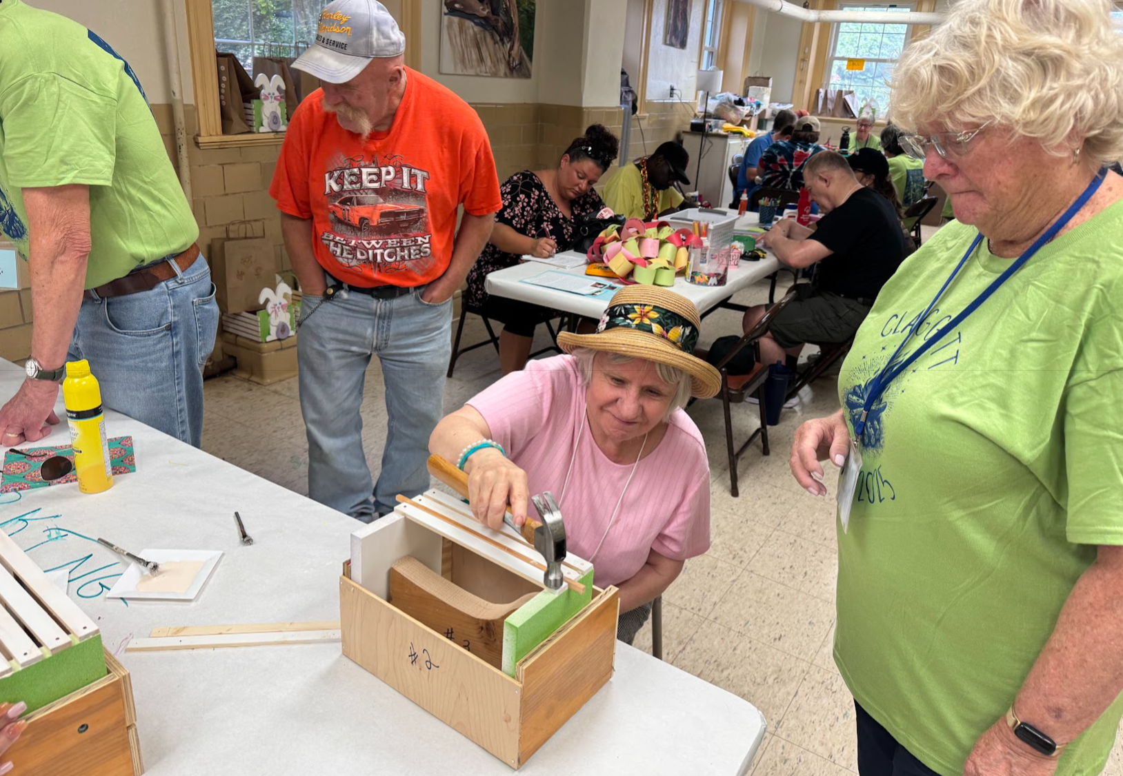 A group of people gathers on grass; two individuals engage in a playful interaction, some seated and observing, under a clear blue sky.