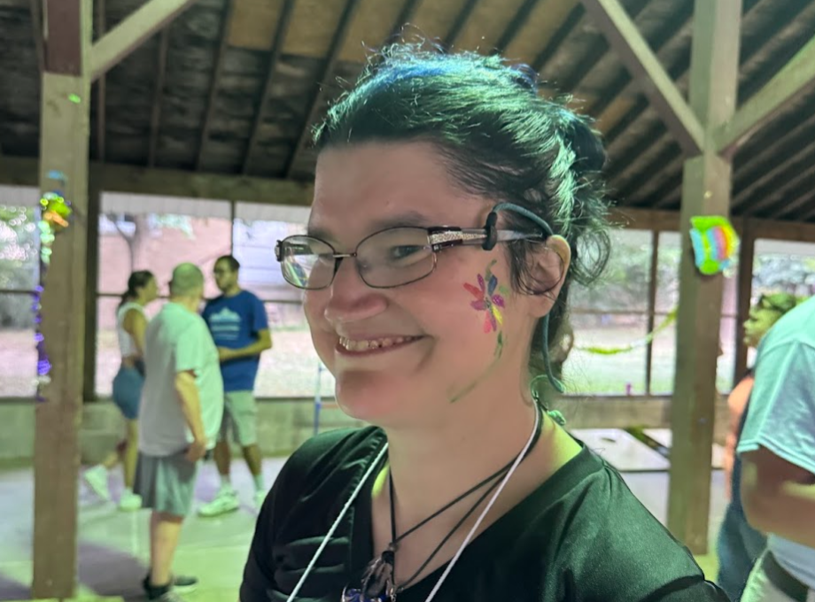 A group of people gathers on grass; two individuals engage in a playful interaction, some seated and observing, under a clear blue sky.