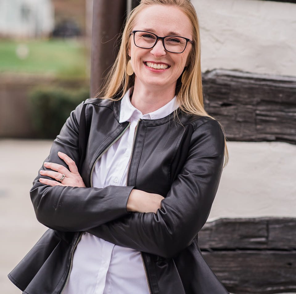 A person smiling outdoors in a green, mountainous landscape with trees and distant hills under a clear sky. No identifiable landmarks visible.